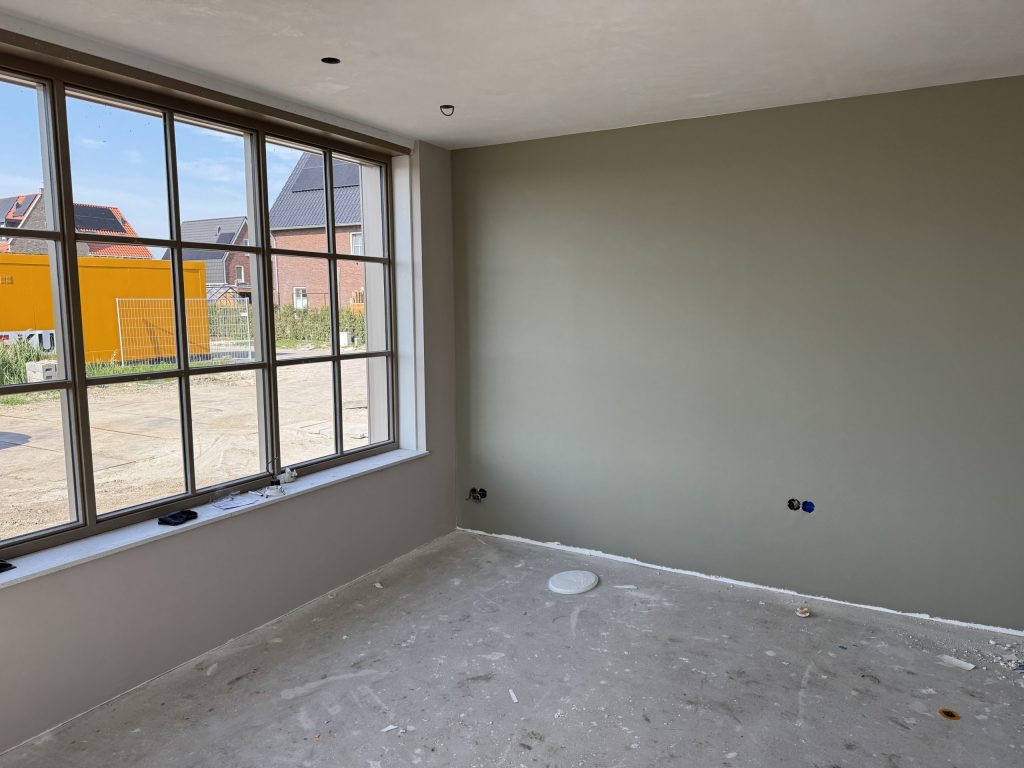Kamer in een nieuwbouwwoning met een egaal afgewerkte grijsgroene wand en een groot raam op de nieuwe straat
